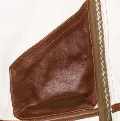 Close-up detail of a brown leather pocket with contrast stitching and a silver zipper, set against a white fabric background.