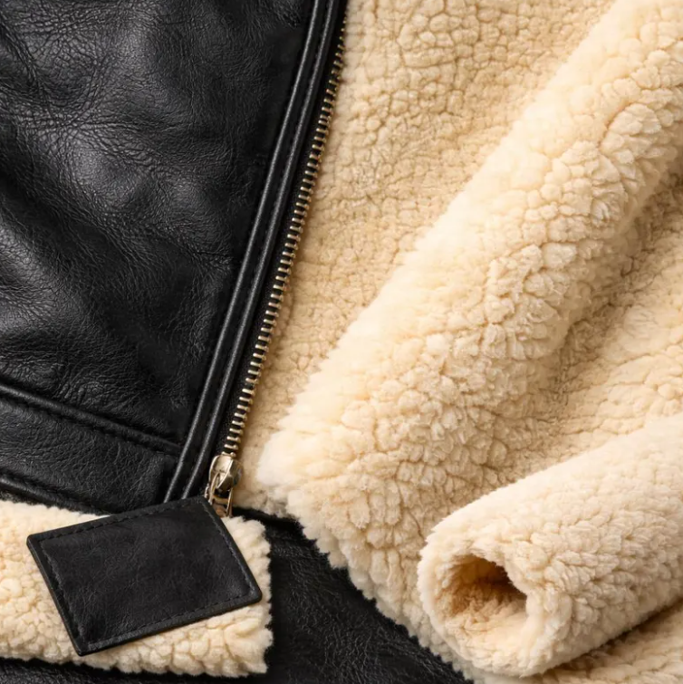 Close-up detail of a jacket showing black leather exterior, a gold-tone zipper, and a thick cream sherpa fleece lining.